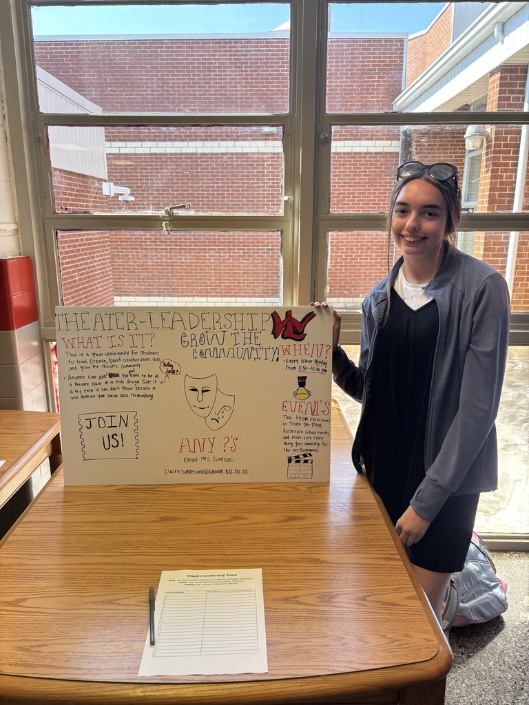 A person stands behind a table, holding a poster with text and a drawing. The table is wooden, with a pen and paper on it. A window with a brick building in the background is behind them.
