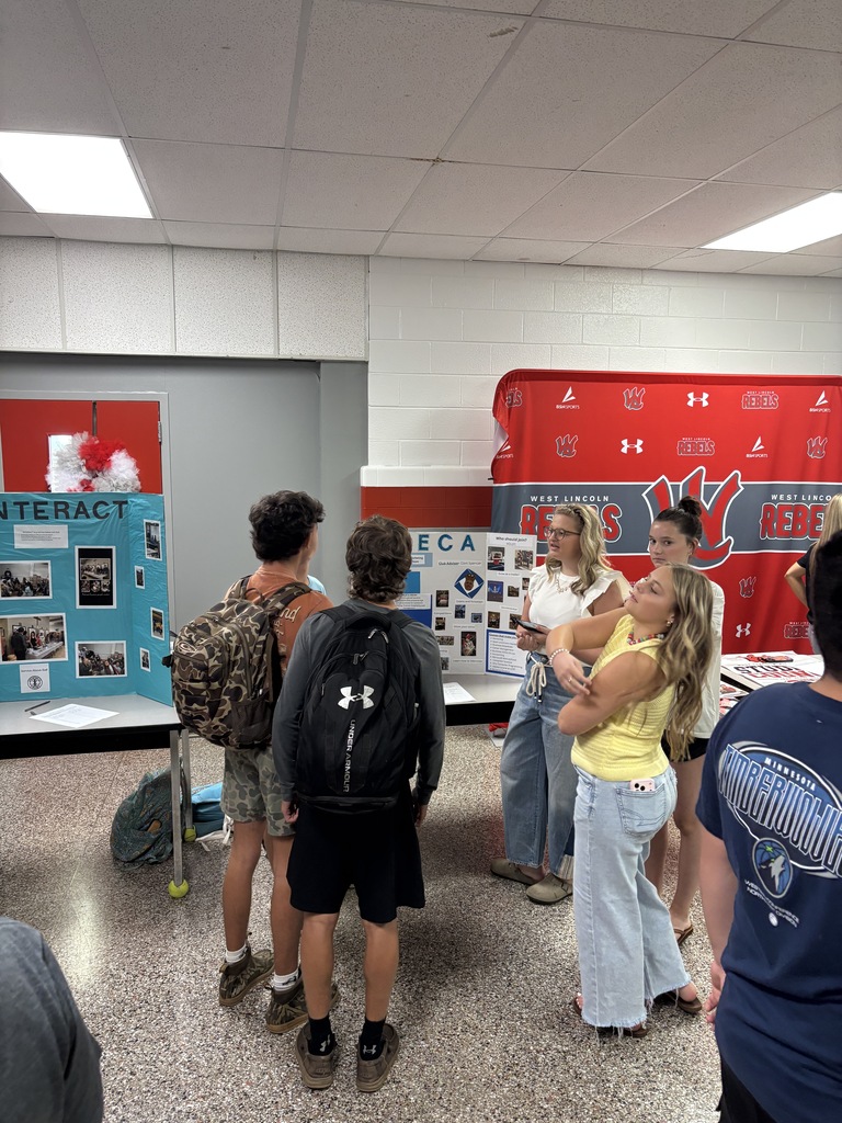 A group of people gather near a red and white backdrop in a room. Some wear backpacks.