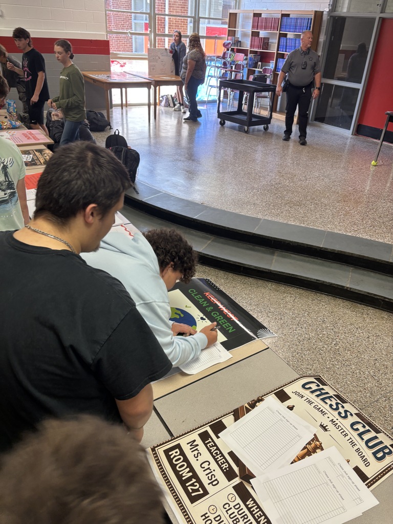 Students are gathered in a room with tables and chairs. Some are seated, while others are standing. A person writes at a table.