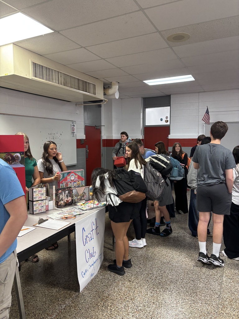 Multiple people stand in a line, possibly at a school event, with tables and various items on display.