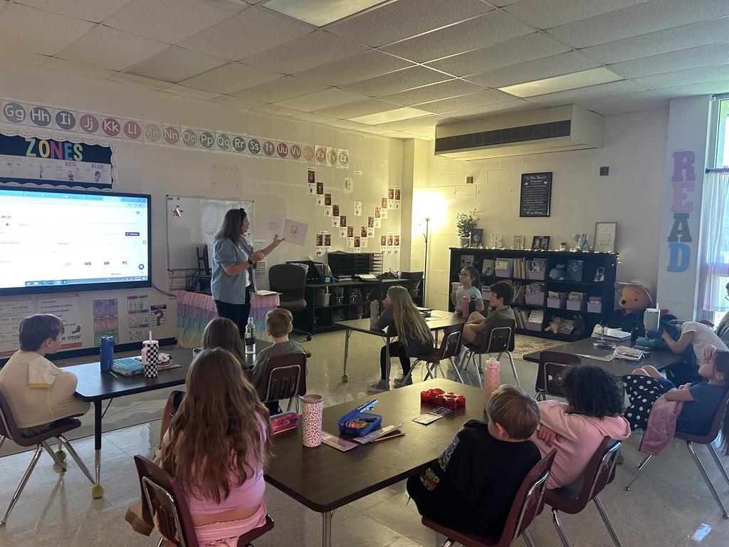 The principal stands at the front of a brightly lit elementary classroom holding an open children's book, reading to a class of students seated around small group tables listening attentively.