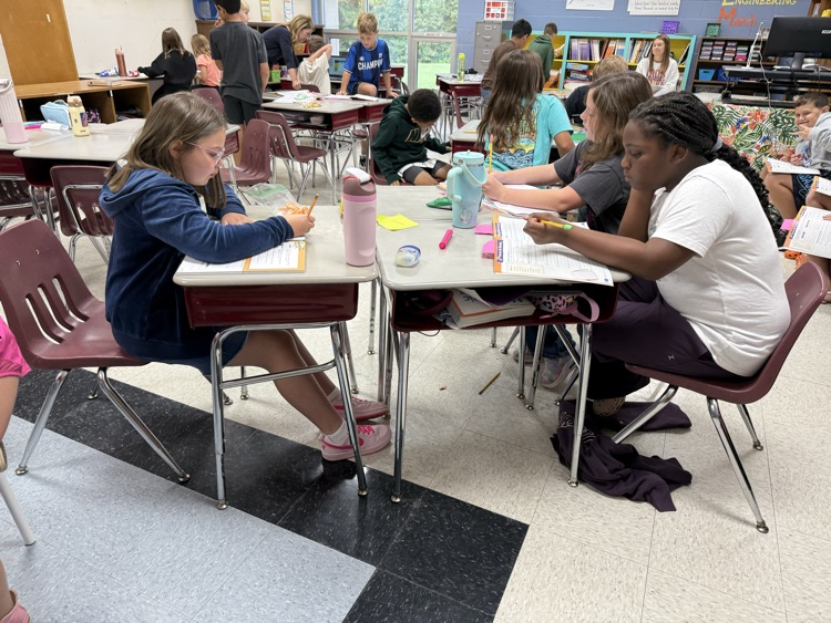 Elementary students sit at pushed-together desks in a classroom, focused on writing and working independently in their workbooks during a lesson.