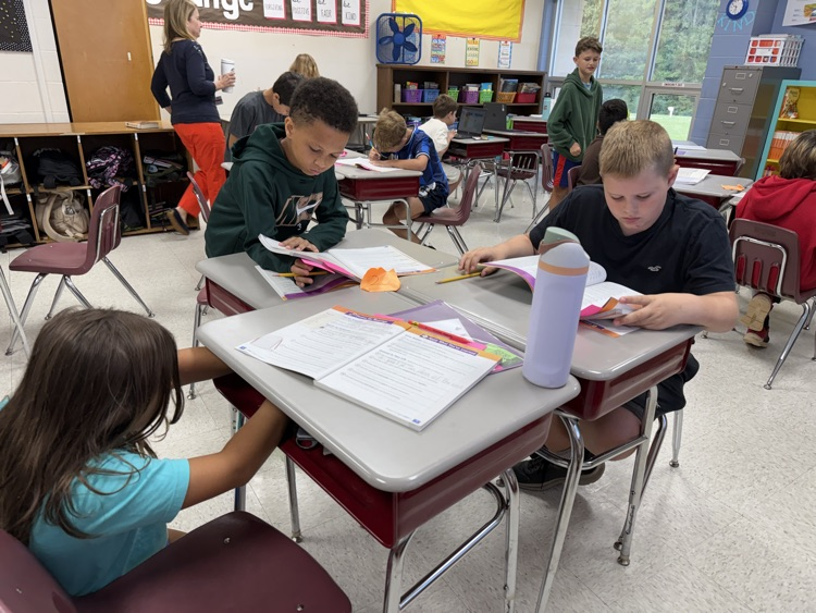 Elementary students seated at a cluster of desks in a classroom setting, reading open workbooks and completing written assignments.