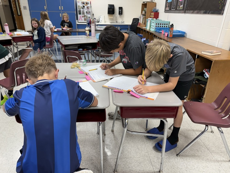 Three elementary students lean over a small group of desks in a classroom, actively writing and working together in open workbooks.