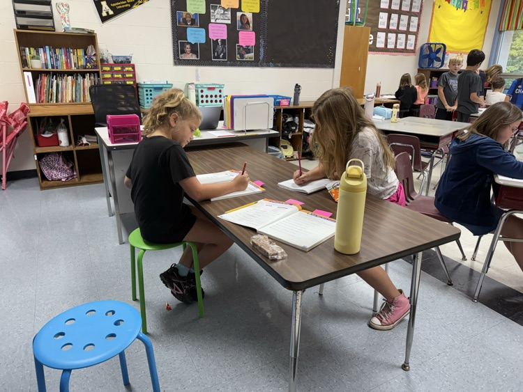 Two elementary students sit across from each other at a long wooden table in a classroom, quietly writing in open workbooks with pencils.