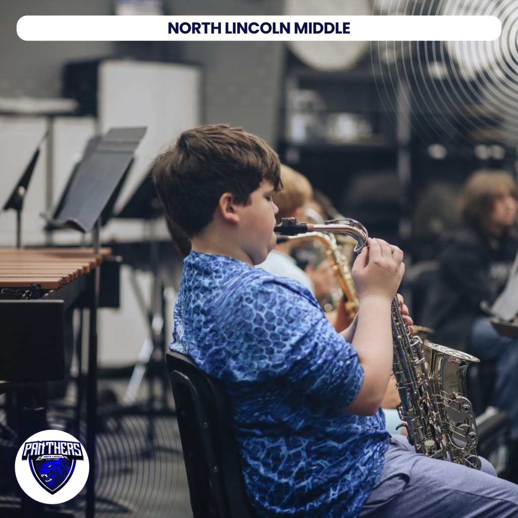 A young boy plays a saxophone while seated in a classroom, with musical instruments and other students in the background.