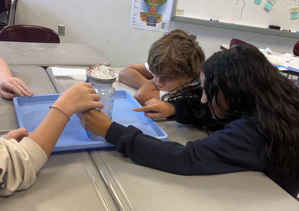 Two students in a classroom, one holding a clear plastic container, both examining an object inside.