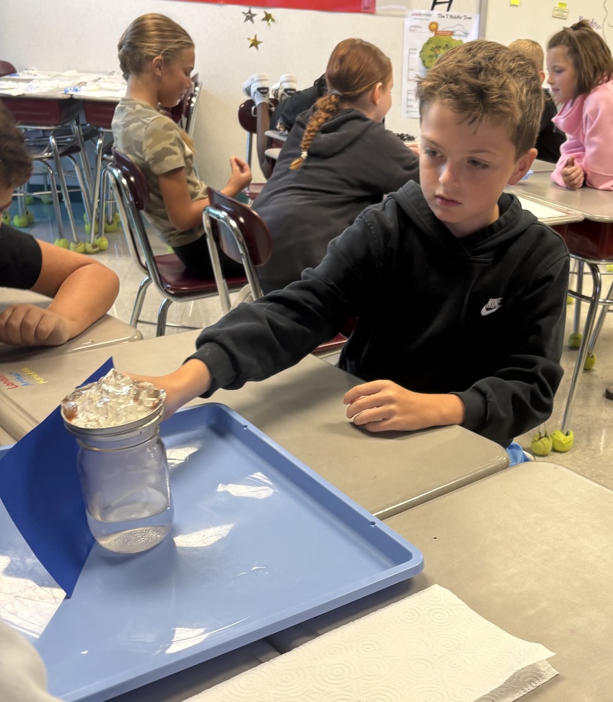 A boy in a black hoodie is seated at a table, handling a jar filled with ice.