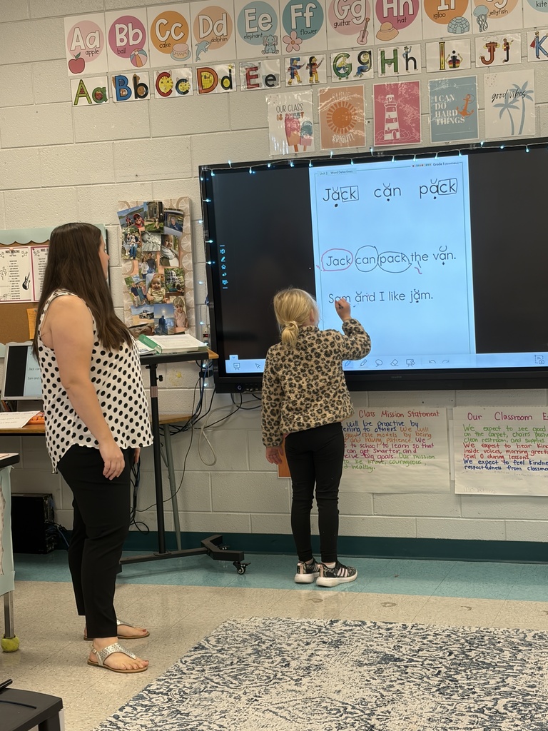 Two people, one standing and one seated, are in front of a large screen. The screen displays text.