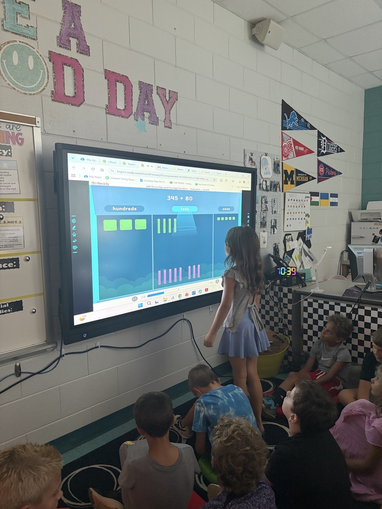 A group of children watch a presentation on a large screen in a classroom. The screen displays a graph.