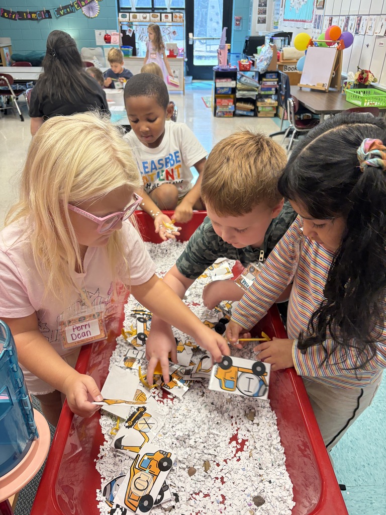 Four children examine objects in a red tray filled with snow. A table, balloons, and other children are in the background.