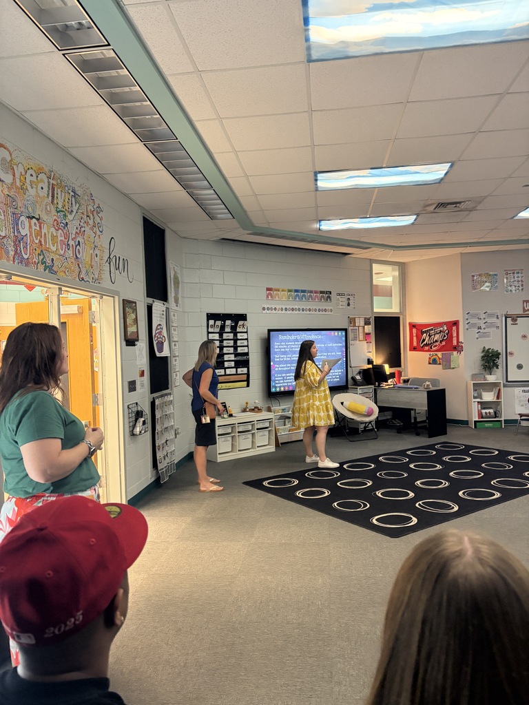 A classroom with four people. A woman stands in front of a television, while others look on.