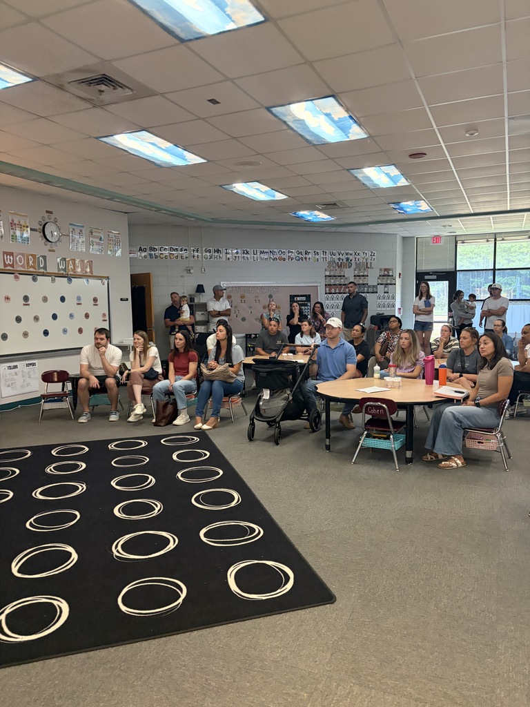 A group of people sitting in a classroom with a whiteboard in the background and round floor mat.