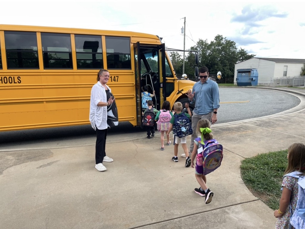 People are walking to a school bus on the curb. A man and a woman stand near the bus.