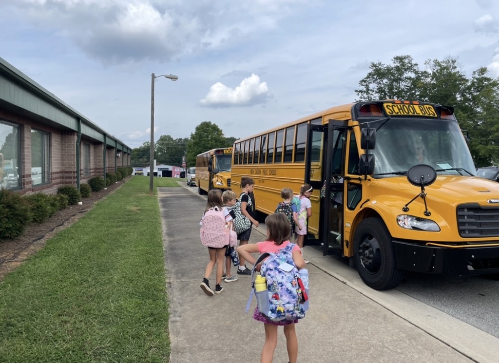 Children wearing backpacks walk towards a yellow school bus. Buildings and green lawn in background.