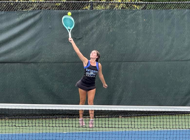 A woman on a tennis court, mid-swing, holding a tennis racket, reaching for an airborne yellow ball.