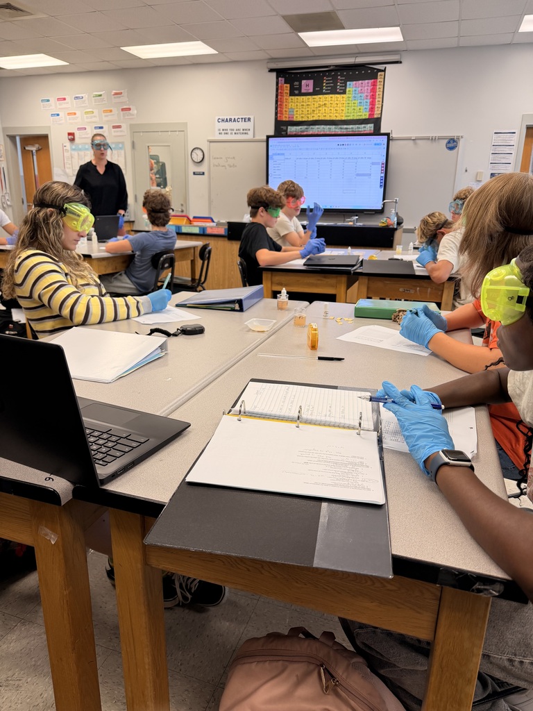Students wearing goggles and gloves work in a classroom with a large screen and tables.