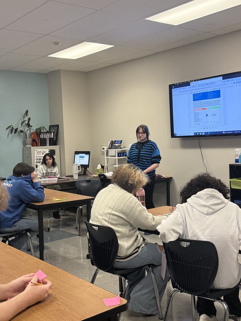 A classroom with a teacher at a desk in front of a screen, and students sitting at tables.
