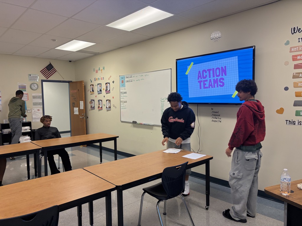 Four people stand in a classroom with a whiteboard and monitor. One person reads a paper at a table.