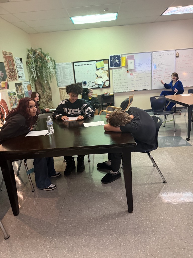 A classroom with students seated around a desk, one with head down. Two seated on chairs, one with paper. A teacher behind a desk. Whiteboard and bulletin boards in background.