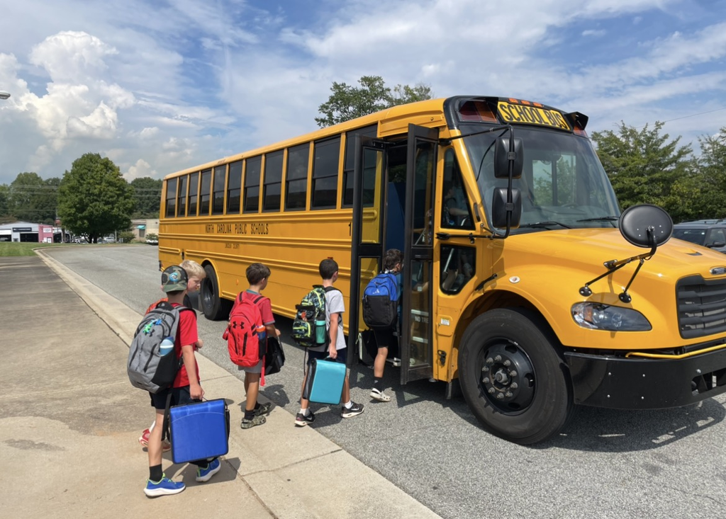 Three kids with backpacks and bags enter a yellow school bus on a street with trees and clouds.