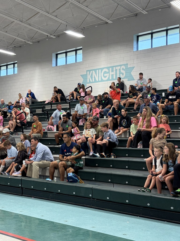 Many people sitting on green bleachers in a school gym, with a sign reading "Knights" on the wall.