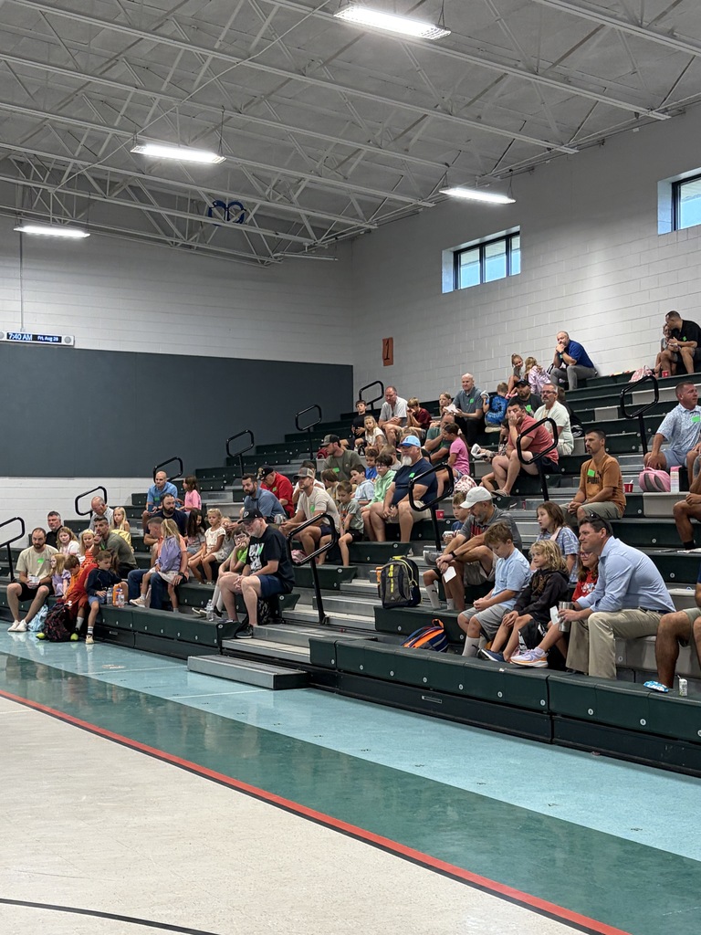 A large group of people are seated in an indoor auditorium. The room has high ceilings, windows, and rows of benches.
