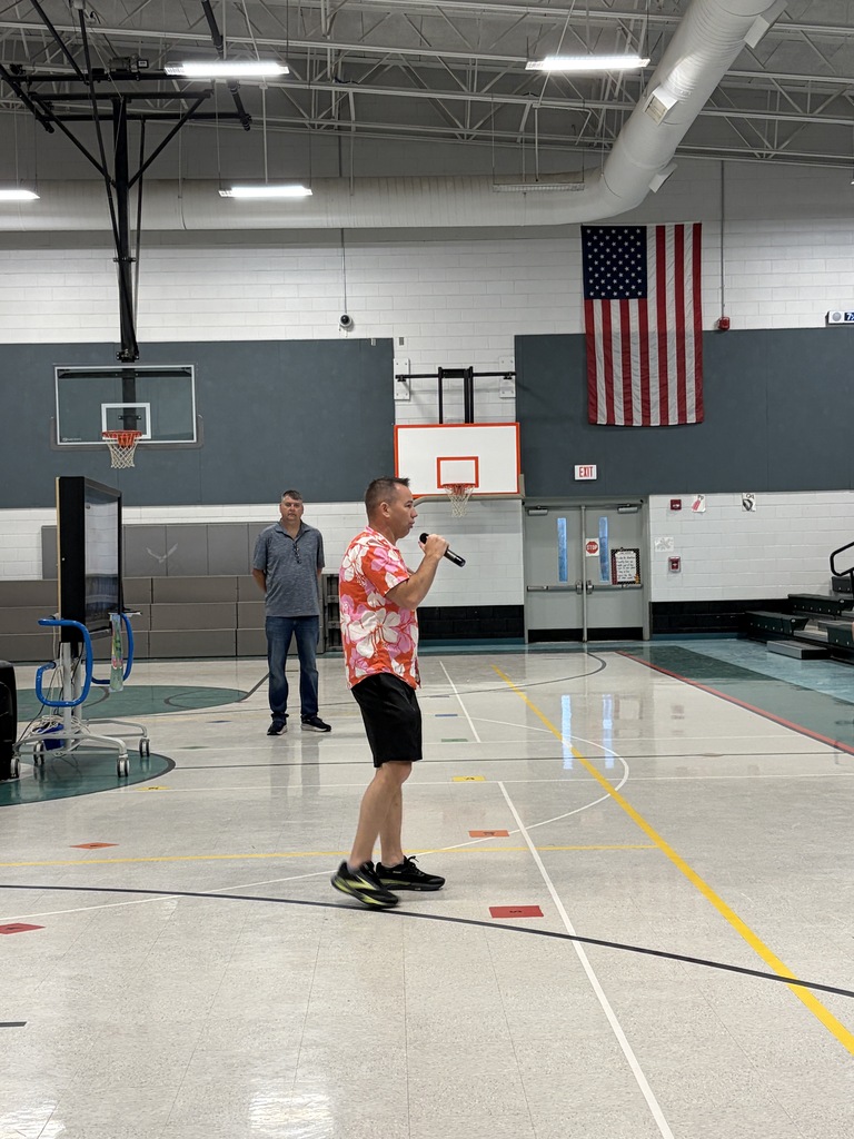 A person holding a microphone stands in an indoor basketball court with a flag on the wall.