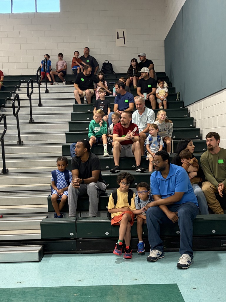 Spectators in a gymnasium watch a group of people sit on bleachers, including adults and children.