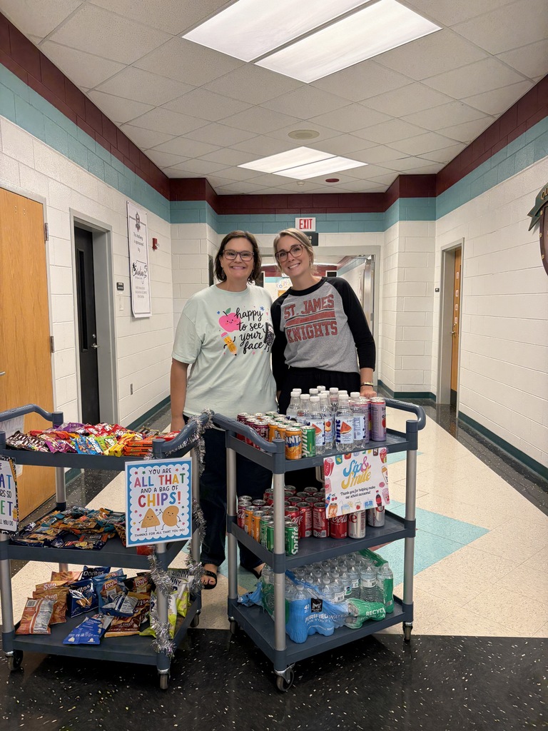 Two people stand next to a food cart and a drinks cart in a school hallway.