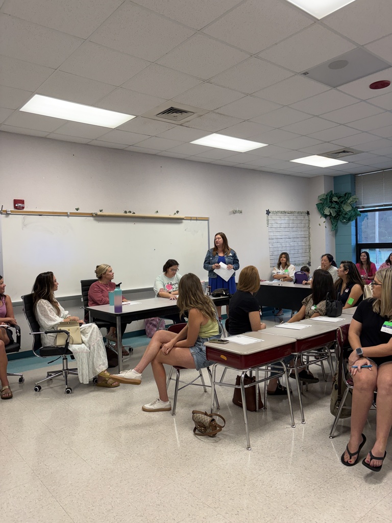 A classroom with people sitting in chairs around desks, listening to a standing woman who appears to be speaking.