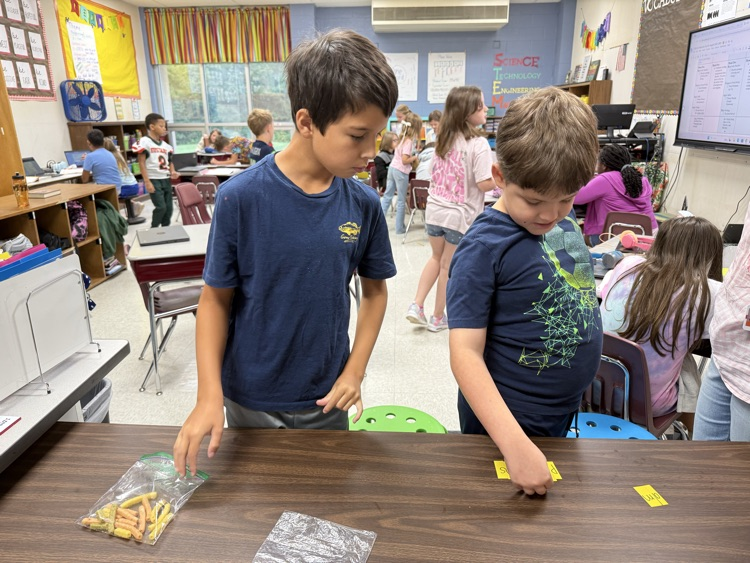 Two elementary students collaborate at a table during a classroom activity, focusing on arranging small yellow word strips alongside a bag of colorful learning manipulatives.