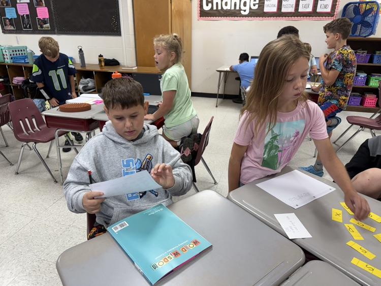 Elementary students participating in classroom learning activities. In the foreground, one student seated at a desk reads a paper with a teal Word Wisdom workbook nearby, while another student stands beside the desk arranging small yellow letter strips.