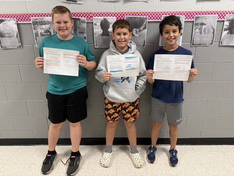 Three elementary students stand side by side in a hallway smiling while holding up completed worksheets with highlighted text and top scores.