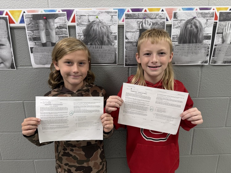 Two elementary students stand side-by-side in front of a classroom display, smiling as they hold up completed reading comprehension worksheets marked with top scores.