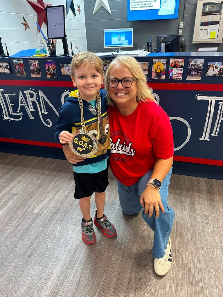 A woman and a young boy stand together in a room with a blue wall. The boy has a gold medal around his neck.