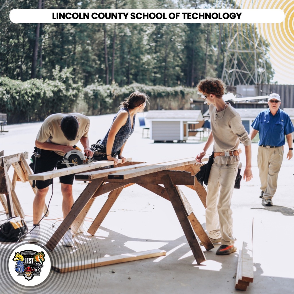Four people working on a wooden table outdoors. One person uses a power saw. A logo is at the bottom left.