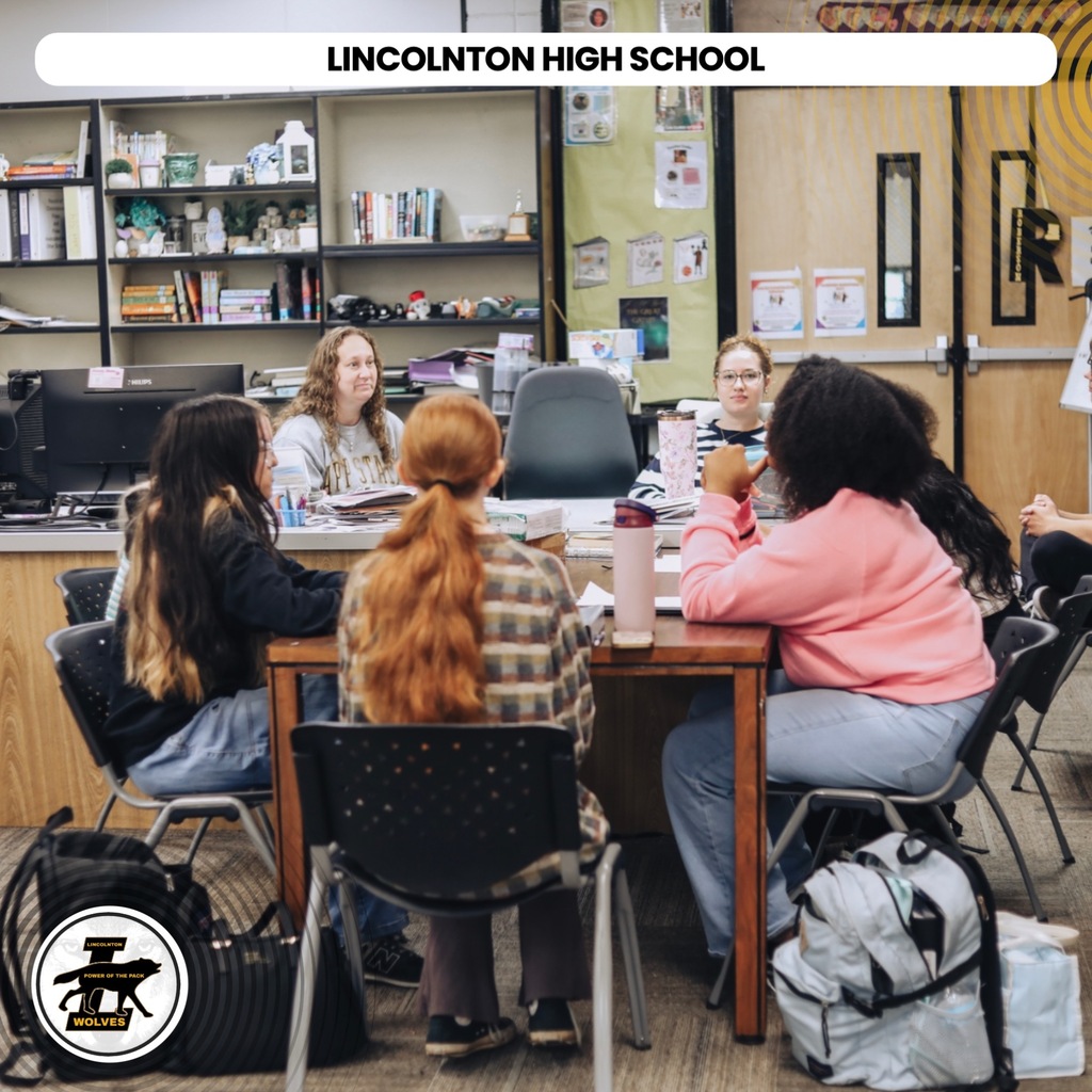 Several students sit at a table in a classroom with shelves and a door in the background.