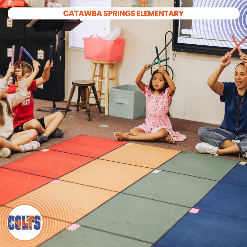 Several children hold instruments. A teacher smiles at them. A classroom is in the background.