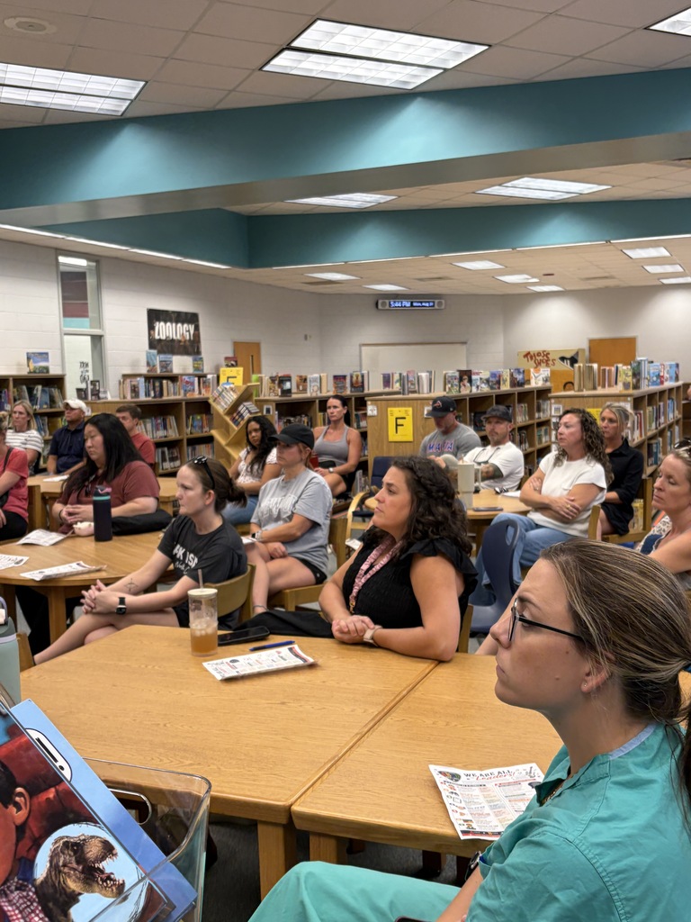 A group of people are seated around tables in a library. Some wear glasses and watches, and others hold books.