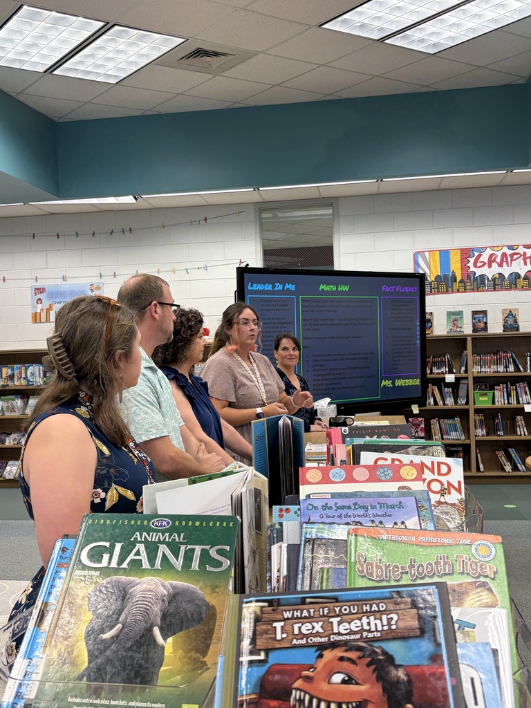 A group of people standing and sitting around a monitor with books on a table in a library.
