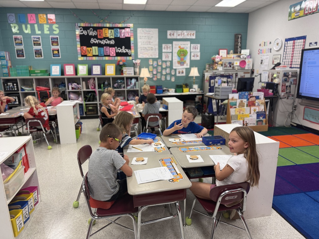 Children seated at desks in a classroom with a colorful rug and a bulletin board.