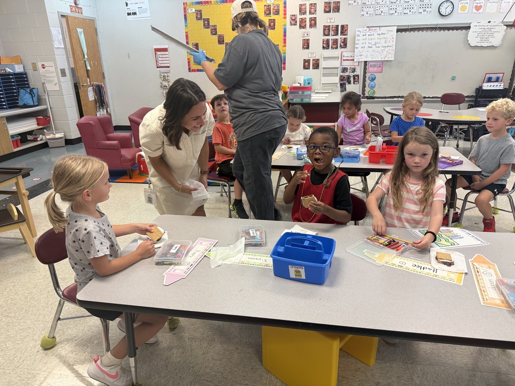 Several young children, some seated at tables, are engaged in an art activity. An adult stands behind them, offering assistance.