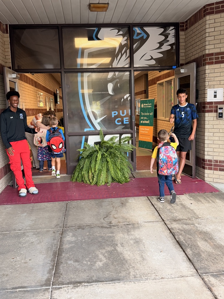 Several children wearing backpacks enter a school building with a man and another adult.