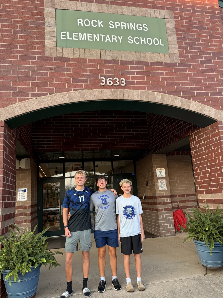 Three young individuals stand in front of a brick building. The sign reads "ROCK SPRINGS ELEMENTARY SCHOOL 3633." Two boys wear sneakers, and one wears a watch. Potted plants flank the entrance.