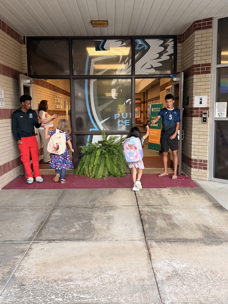 Five children with backpacks enter a building, two adults stand nearby. Brick walls, a glass door with an eagle logo.