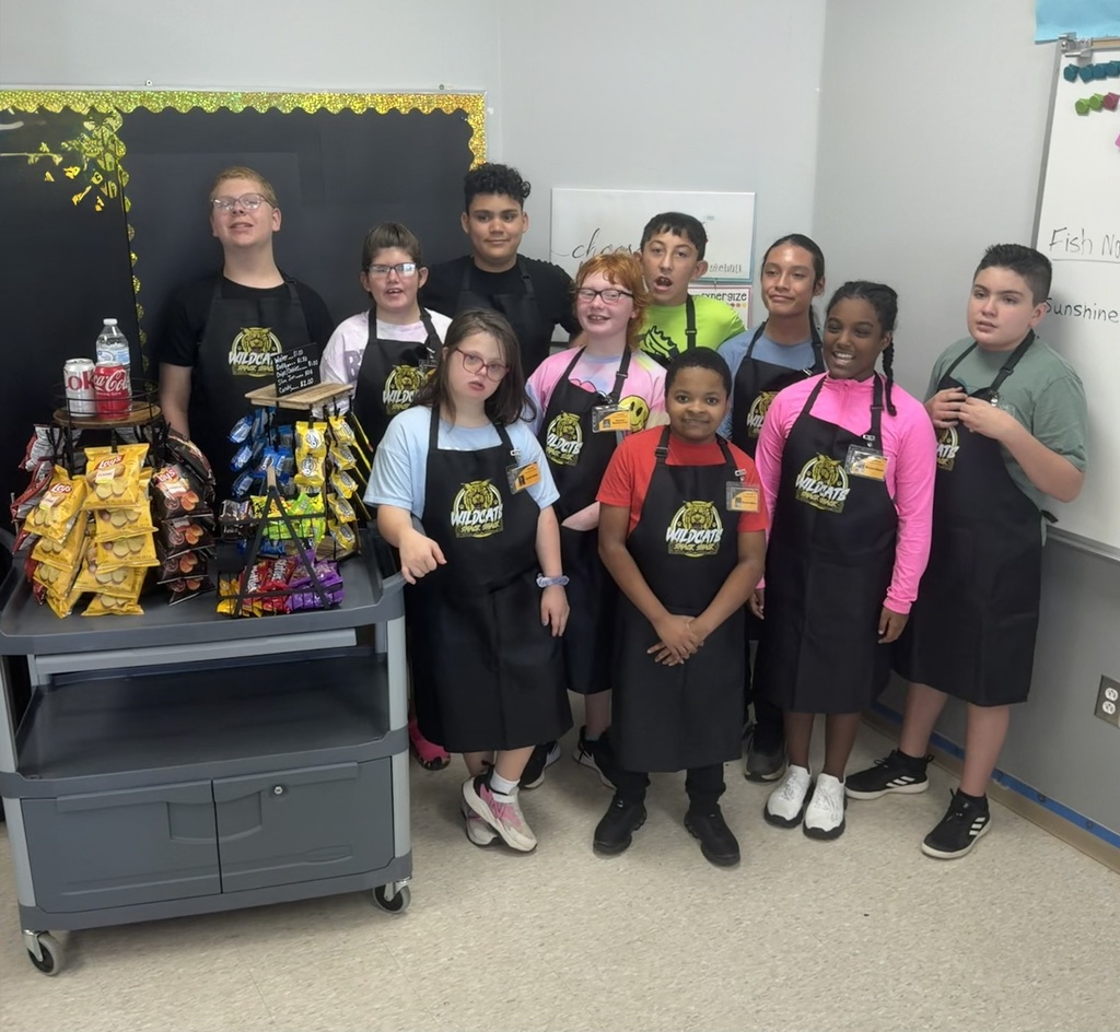 A group of children in black aprons pose near a cart with snacks in a classroom.