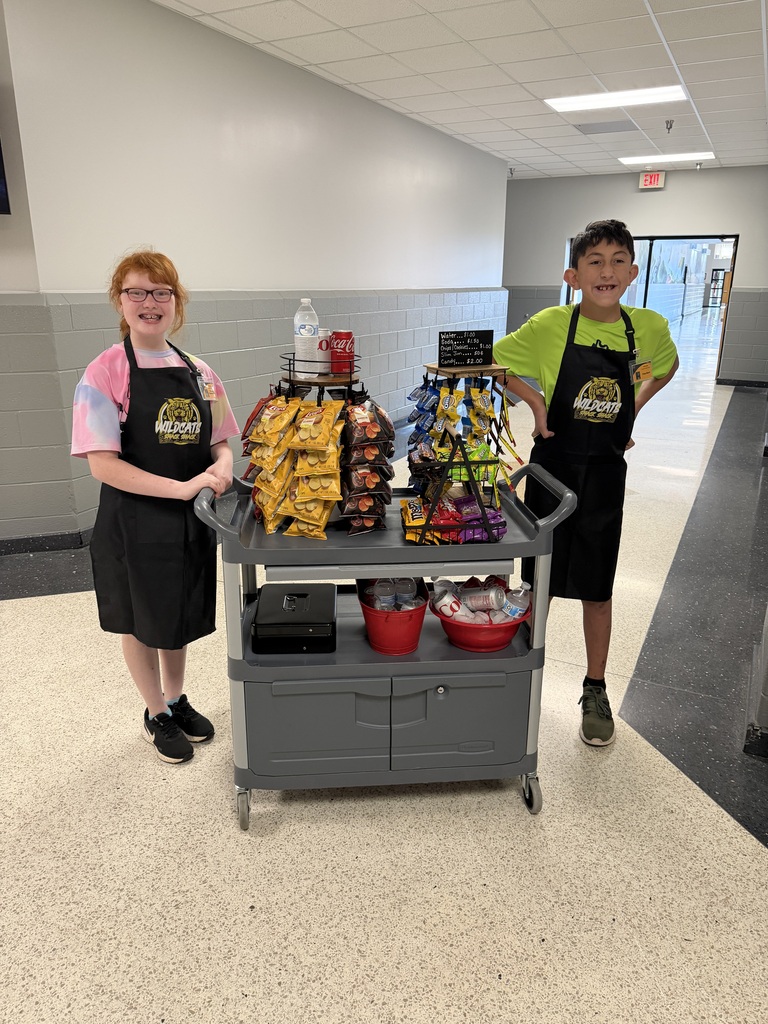 Two children in aprons, standing behind a cart filled with snacks, in a bright hallway.