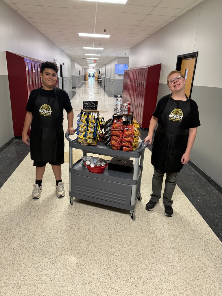 Two individuals stand beside a cart filled with snacks in a school corridor with white flooring and red lockers.