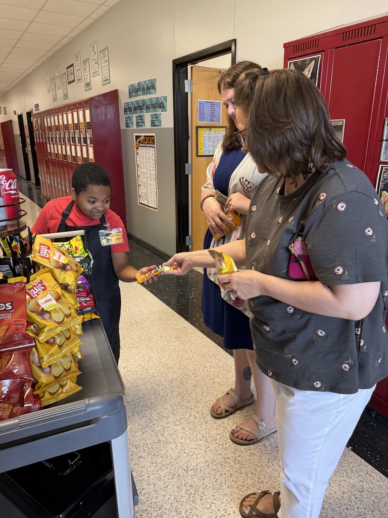 Three adults near a table with snacks. A boy in an apron holds a bag. Behind them, lockers line the walls.